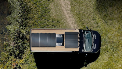 Aerial view above a van with solar panels on the roof. They are parked in a remote area with brush near the trail. Between the panels, a rooftop air conditioner provides comfort.